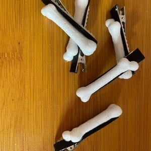 May include: Four hair clips with a bone design. Each clip has a black base and a white, glittery bone shape. The clips are arranged on a wooden surface.