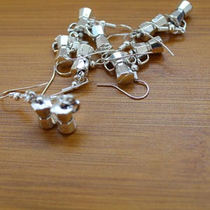 May include: Silver-toned coffee pot earrings with a hook closure. The earrings are small, detailed replicas of a stovetop espresso maker. They are scattered on a wooden surface, showcasing their design and craftsmanship.