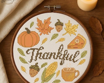 May include: A cross-stitch embroidery hoop with a fall theme. The design includes a pumpkin, leaves, acorns, corn, a slice of pie, and a mug of coffee. The word "Thankful" is stitched in the center. A lit candle is in the background.