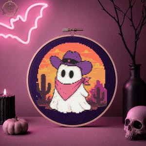 Puede incluir: Un bastidor de bordado circular con un patrón de punto de cruz de un fantasma blanco con un sombrero de vaquero morado y un pañuelo rosa. El fondo muestra un paisaje desértico con cactus bajo una puesta de sol naranja sobre tela oscura.