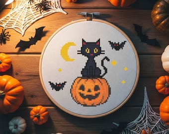 May include: A cross-stitch embroidery hoop featuring a black cat sitting on a jack-o'-lantern, with a yellow crescent moon and bats. The Halloween-themed design is surrounded by miniature pumpkins, spiderwebs, and bats on a wooden surface.