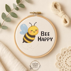 Puede incluir: Un bastidor de bordado de punto de cruz con una abeja amarilla y negra sonriente con alas azules. El texto "BEE HAPPY" está bordado en negro. El bastidor está sobre un fondo neutro con eucalipto e hilo.