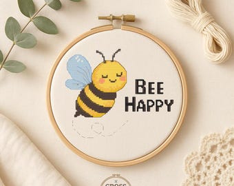 Può includere: Un telaio da ricamo a punto croce con un'ape gialla e nera sorridente con ali blu. Il testo "BEE HAPPY" è ricamato in nero. Il telaio è ambientato su uno sfondo neutro con eucalipto e filo.