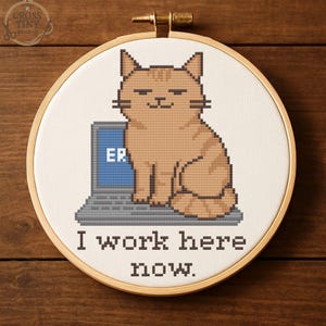 Könnte beinhalten: Kreuzstich-Stickerei in einem Holzreifen. Sie zeigt eine pixelige, orangefarbene Katze auf einem Laptop mit "ER" auf dem Bildschirm. Der Text darunter lautet "I work here now". Das Design befindet sich auf weißem Stoff, umrahmt von einem Holzreifen.