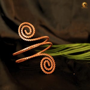 May include: A copper arm cuff with a spiral design. The cuff wraps around the arm with two spiral accents at each end. The metal has a twisted rope texture. The background is dark with a hint of green.