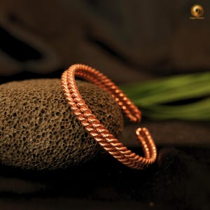 Op de afbeelding: Een metalen koperen manchetarmband met een dubbelstrengs gedraaid touwontwerp. De armband heeft een open constructie en een gepolijst, reflecterend oppervlak dat het licht opvangt en de textuur van de gedraaide metaaldraad laat zien.