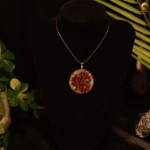 Op de afbeelding: Een ronde hangerketting met drie donkerrode koperdraadspiralen ingebed in een schijf van heldere hars. De hars bevat kleine, veelkleurige steenfragmenten in groen-, blauw- en goudtinten. De hanger hangt aan een dun zwart koord.