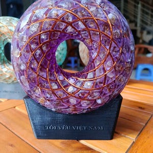 May include: A spherical, amethyst-colored sculpture with a copper wire lattice design. The sculpture is mounted on a black base with text. The artwork is displayed on a wooden surface.
