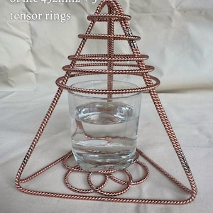 May include: A copper wire pyramid structure featuring five tensor rings and a flower of life base, surrounding a clear glass filled with water. The text 1 pyramid + 1 flower of life 432mhz + 5 tensor rings is visible in the upper left corner.