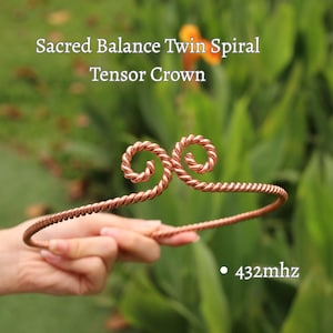 May include: A copper-colored Tensor Crown with a twin spiral design. The crown is held in front of a blurred green background. The text "Sacred Balance Twin Spiral Tensor Crown" and "432mhz" are visible.