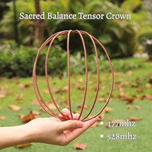 May include: A copper-colored Sacred Balance Tensor Crown is held in a hand. The crown is made of twisted wire and has a spherical shape. Text on the image reads "Sacred Balance Tensor Crown", "177mhz", and "528mhz". The background is blurred green foliage.