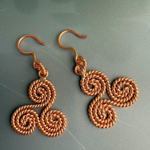 May include: A pair of copper-colored earrings featuring a triskelion design made from twisted wire. Each earring consists of three interconnected spiral shapes attached to a simple curved ear wire hook, showcasing a metallic finish.