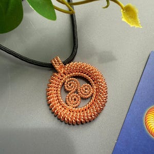 Op de afbeelding: Koperkleurige hangerketting met een cirkelvormig ontwerp en een drievoudig spiraalsymbool. De hanger is gemaakt van geweven draad en hangt aan een zwart koord. De ketting wordt gepresenteerd tegen een grijze achtergrond met groen gebladerte.