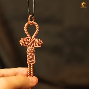 May include: A handcrafted copper ankh pendant on a black cord. The ankh, a symbol of life, is intricately woven from copper wire, showcasing a detailed design. The pendant is held against a blurred dark background.
