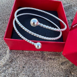 May include: A silver-toned spiral bangle bracelet featuring a twisted rope-like texture throughout the band. The bracelet ends in two smooth, polished spherical balls at each terminal point, resting inside a square red gift box with black lining.