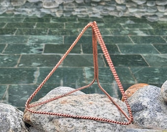 Puede incluir: Una escultura piramidal tridimensional de color cobre con un diseño de cuerda trenzada. La pirámide descansa sobre una gran roca gris. El fondo presenta una fuente de agua con escalones de baldosas verdes y otras rocas.