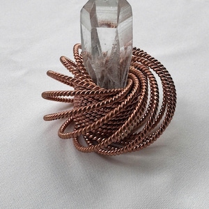 May include: A clear, pointed quartz crystal set within a base of coiled, twisted copper wire. The metallic wire forms a dense, circular nest around the base of the transparent, faceted stone, creating a contrast between the smooth crystal and the textured metal.