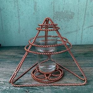 Puede incluir: Un portavelas de metal color cobre con un diseño geométrico. El soporte presenta una base triangular y una serie de círculos concéntricos alrededor de un vaso pequeño. El vaso está diseñado para contener una vela. El metal tiene una textura trenzada.