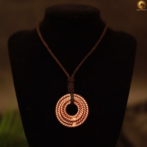 May include: A copper pendant necklace with a circular design. The pendant is made of tightly wound copper wire, creating a layered effect. The necklace has a brown cord and is displayed on a black mannequin.