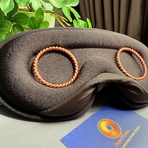 May include: A black sleep mask with two copper-colored circular accents. The mask has a curved shape and a soft, padded appearance. A business card with a logo and text is visible in the foreground.