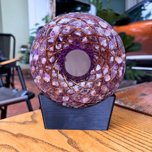 May include: A decorative, spherical object with a central hole, crafted from purple and clear stones, encased in a copper wire lattice. It sits on a black, rectangular base, showcasing a unique blend of materials and design.