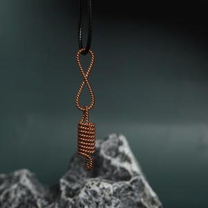 Op de afbeelding: Een koperkleurige hangerketting met een uniek ontwerp. De hanger heeft een oneindigheidssymboolvorm boven een opgerold, cilindrisch element. De ketting hangt aan een zwart koord tegen een donkergroene achtergrond.