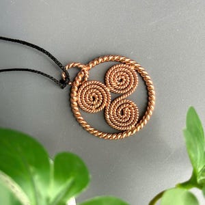 May include: A copper-colored pendant featuring a circular frame made of twisted wire. Inside the circle, three smaller, tightly coiled spirals are arranged in a triangular pattern. The pendant is attached to a thin black cord for wearing as a necklace.