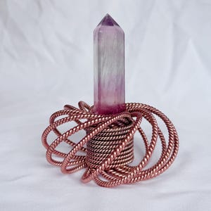 May include: A polished, hexagonal amethyst crystal point with purple and clear banding, set vertically into a base constructed from multiple interconnected loops of twisted, metallic copper-colored wire arranged in a circular pattern.