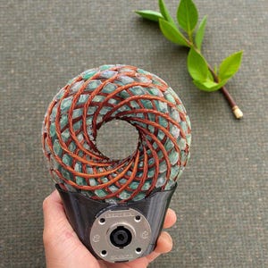May include: A unique, handcrafted sculpture featuring a donut-shaped core of green stones woven with copper wire. The base is black with a silver-colored connector. A sprig of green leaves adds a natural accent.