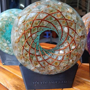 May include: A clear glass sculpture with a spiral design, featuring a central opening. The sculpture is supported by a black base with text. The glass is filled with small, amber-colored pieces and copper wire.