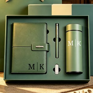 May include: A sage green gift set featuring a notebook with a metal clasp, a ballpoint pen, a small USB drive, and two cylindrical vacuum-insulated tumblers. Each item is marked with the monogram M|K in a clean, white, or dark sans-serif font.