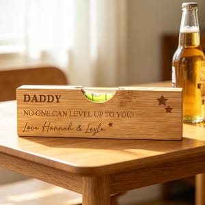 Può includere: Una livella a bolla rettangolare in bambù con il testo inciso DADDY, NO ONE CAN LEVEL UP TO YOU!, e Love Hannah & Layla, insieme a due piccole stelle. L'attrezzo in legno presenta una fiala centrale con liquido verde su un tavolo.