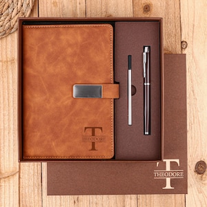 May include: A brown leather journal with a metal clasp and the name "THEODORE" engraved on the cover, presented in a gift box with a pen and refill. The set is displayed on a wooden surface.