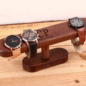 Puede incluir: Un soporte de madera marrón oscuro para relojes con tres relojes de pulsera. El soporte tiene grabados "CP CHRIS POTTER" y "HUMU". Los relojes tienen correas de cuero negro y oro rosa, negro y dorado, y negro y marrón.
