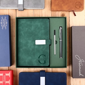 Puede incluir: Un juego de diario y bolígrafo de cuero verde en una caja de regalo, con un cierre plateado. El diario está grabado con la letra "G" y el nombre "Grayson". También se ven otras cajas de regalo y diarios en varios colores.