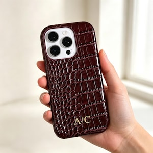 Puede incluir: Una funda para smartphone de color burdeos intenso con una textura brillante de piel de cocodrilo. La funda presenta un recorte para la cámara de triple lente y las iniciales A|C en color dorado grabadas en la parte inferior central.