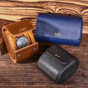 May include: Three leather watch cases in brown, blue, and black, each with a personalized monogram. The brown case is open, displaying a watch with a black face and red strap. The cases are on a wooden surface.