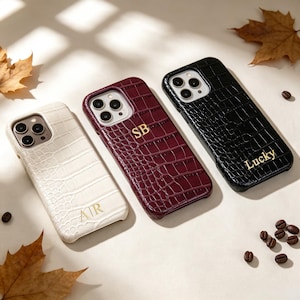 Puede incluir: Tres fundas para smartphone con textura de piel de cocodrilo. La funda de la izquierda es crema con las letras AIR, la central es burdeos con las letras SB y la de la derecha es negra con la palabra Lucky impresa en letras doradas.