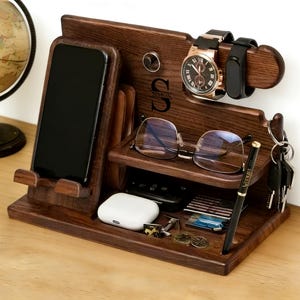 Peut inclure: Une station d'accueil en bois brun foncé avec un support pour téléphone, un porte-montre, une étagère pour lunettes et un crochet pour clés. Le bois est gravé de la lettre S et du nom SIMON. Elle contient un smartphone, deux montres, des lunettes, des clés, un stylo et des écouteurs.