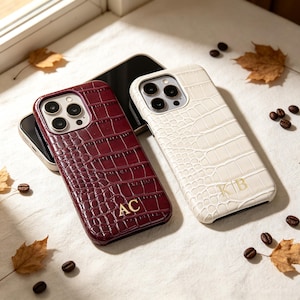 Puede incluir: Dos fundas para smartphone con textura de cocodrilo. Una funda es de color burdeos oscuro con las iniciales doradas AC, y la otra es de color crema con las iniciales doradas KB. Ambas tienen aberturas para un sistema de triple cámara.