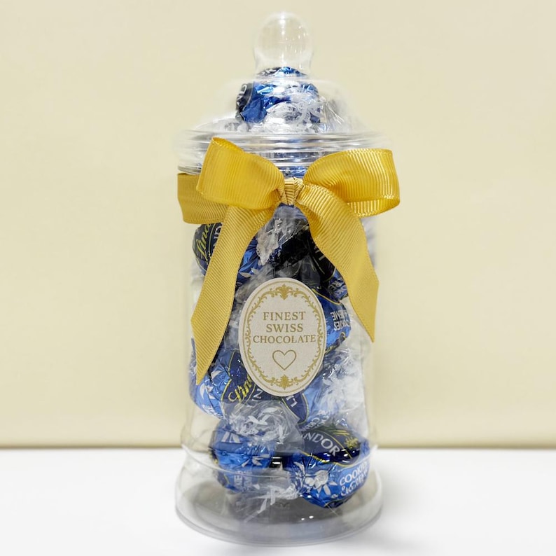 May include: A clear glass apothecary jar filled with blue and silver wrapped chocolates. The jar is tied with a gold ribbon and a label that reads "Finest Swiss Chocolate". The jar has a clear glass lid.