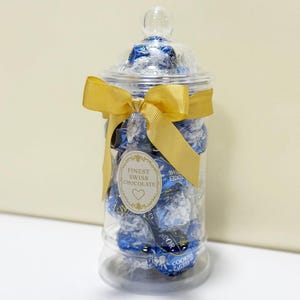 May include: A clear glass jar filled with blue-wrapped chocolates, tied with a gold ribbon. The jar has a clear lid and a label that reads "Finest Swiss Chocolate". The jar is a decorative container for sweets.