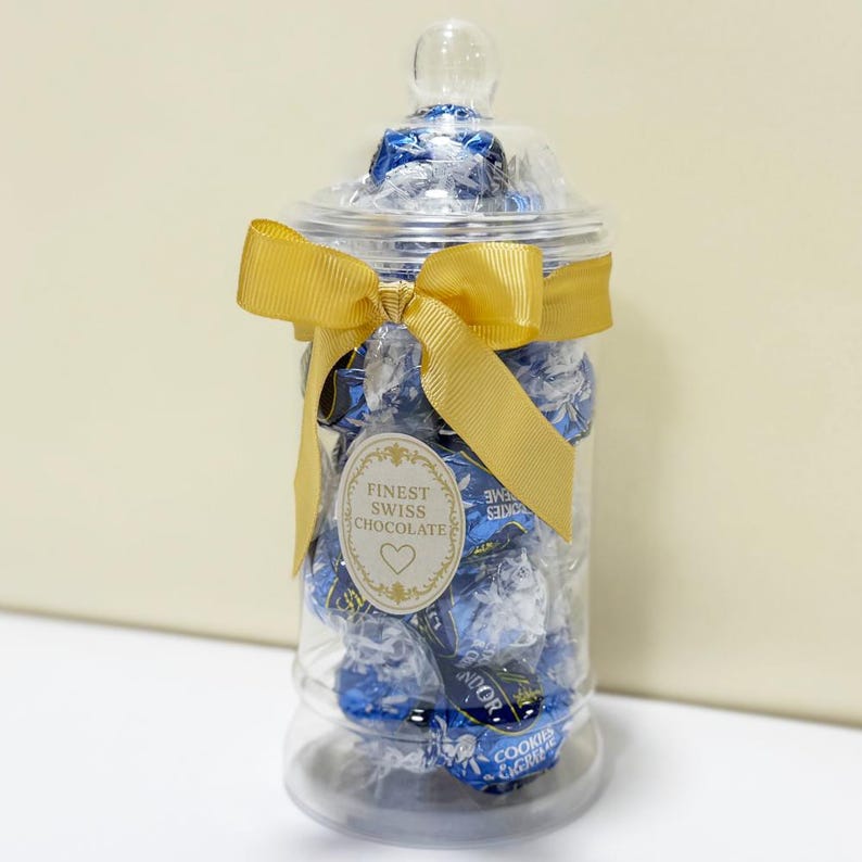 May include: A clear glass jar filled with blue-wrapped chocolates, tied with a gold ribbon. The jar has a clear lid and a label that reads "Finest Swiss Chocolate". The jar is a decorative container for sweets.