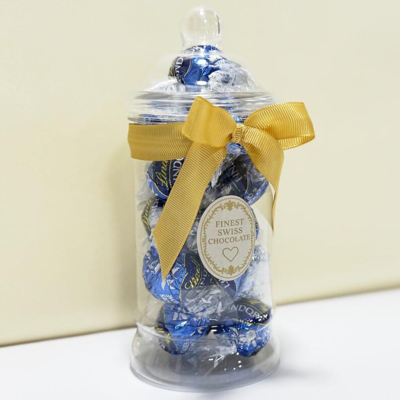 May include: A clear glass jar filled with blue-wrapped chocolate candies. The jar is tied with a gold ribbon and a tag that reads "Finest Swiss Chocolate." The jar has a clear lid and sits on a white surface.