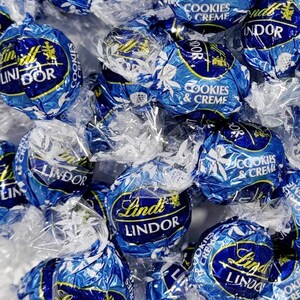 May include: Close-up of several individually wrapped Lindt Lindor Cookies & Creme chocolate truffles. The wrappers are a deep blue with white floral designs and gold accents. The Lindt Lindor logo is prominently displayed on each truffle.