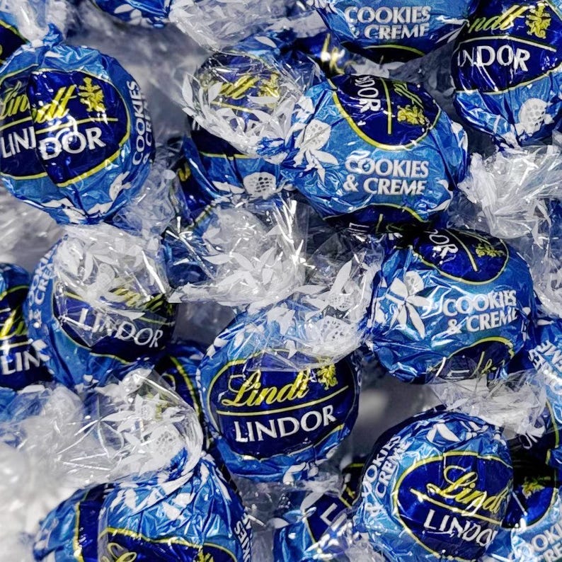 May include: Close-up of several individually wrapped Lindt Lindor Cookies & Creme chocolate truffles. The wrappers are a deep blue with white floral designs and gold accents. The Lindt Lindor logo is prominently displayed on each truffle.