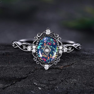 May include: A silver ring with an oval, iridescent gemstone. The gemstone is set in a decorative, black frame with small white stones. The ring band features a twisted design with additional small white stones. The ring is displayed on a dark, textured surface.