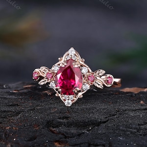 May include: A rose gold ring featuring a pear-shaped red center stone surrounded by small clear accent stones. The band is decorated with metal leaf motifs and additional small red stones on each side, creating a detailed botanical design.