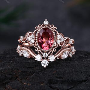 May include: A rose gold ring set featuring a central oval-cut dark pink gemstone surrounded by a decorative filigree frame. The band and the accompanying curved guard ring are adorned with multiple small, clear, round and marquise-cut stones.