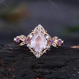 Pode incluir: Um anel em ouro rosa com uma pedra preciosa central rosa claro em forma de pera, rodeada por pequenas pedras transparentes. O aro é decorado com motivos de folhas metálicas e pequenas pedras roxas em cada lado da cravação central.